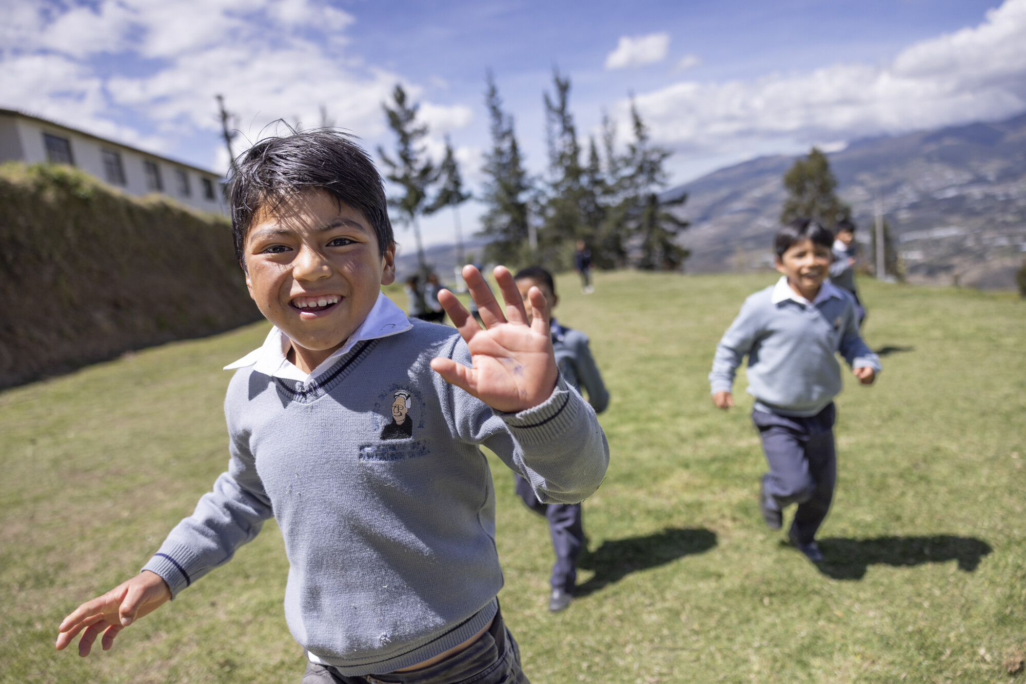 Smiling boy in a school uniform waves while running on a grassy hill, with other children playing behind him and a mountainous landscape in the background.