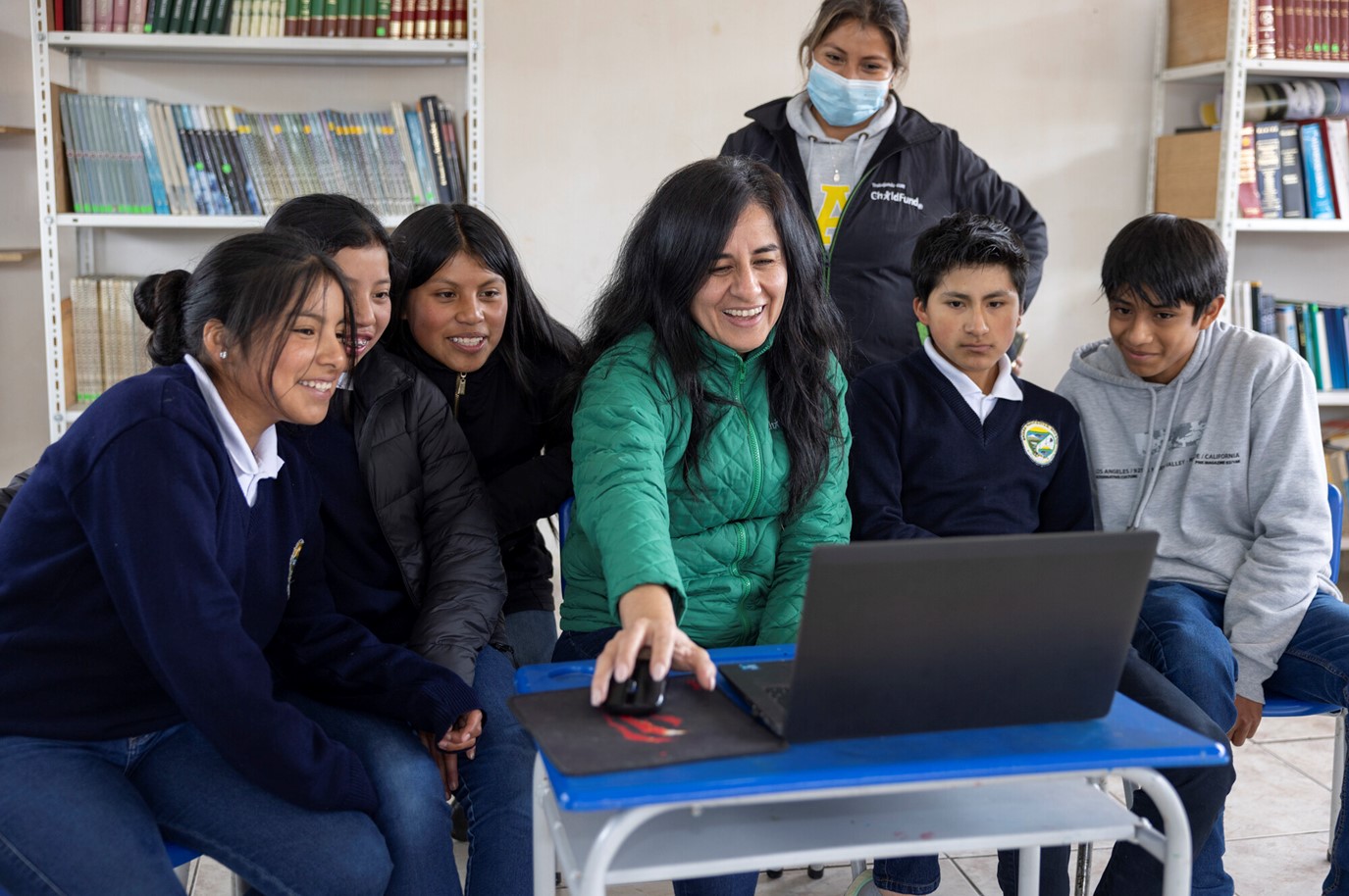 A teacher and a group of students in a classroom smile and engage with a laptop as they participate in a digital safety lesson. A ChildFund staff member observes in the background.