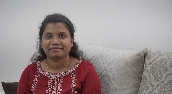 Woman in a red embroidered top sitting on a couch with decorative pillows, looking at the camera.