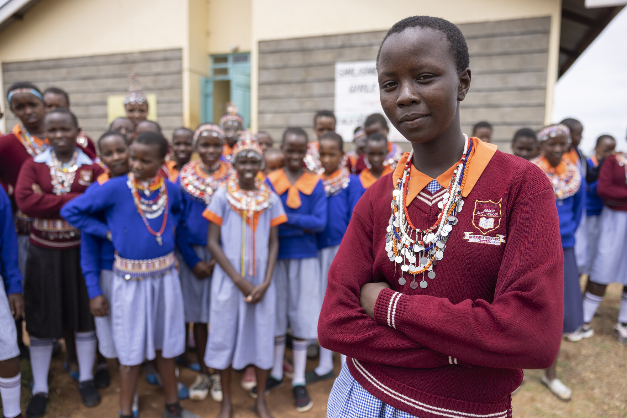 A confident young girl wearing a maroon school uniform and traditional beaded necklaces stands with her arms crossed in front of a group of students outside a school.