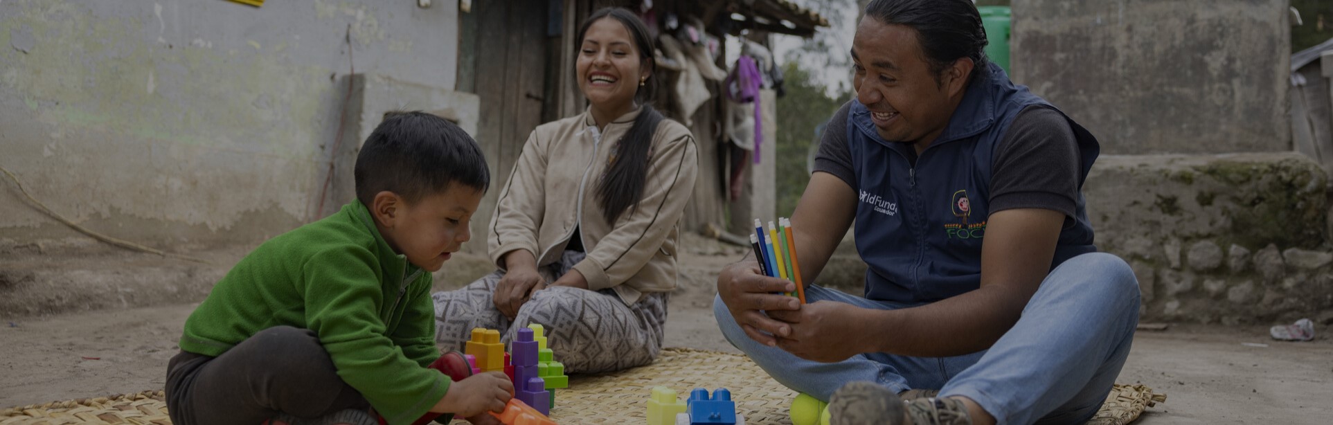 Boy playing with building blocks on a mat, with a smiling woman and man engaging with him in a rustic outdoor setting.