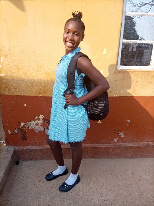 Young girl in Sierra Leone stands wearing backpack and smiling at camera.