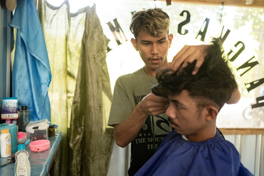 Young barber giving a haircut inside a small shop, with grooming tools on a table and natural light filtering through a curtain.