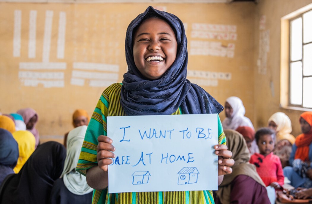 Smiling girl in a headscarf holds a sign reading "I WANT TO BE SAFE AT HOME," standing in a classroom with other girls and women.