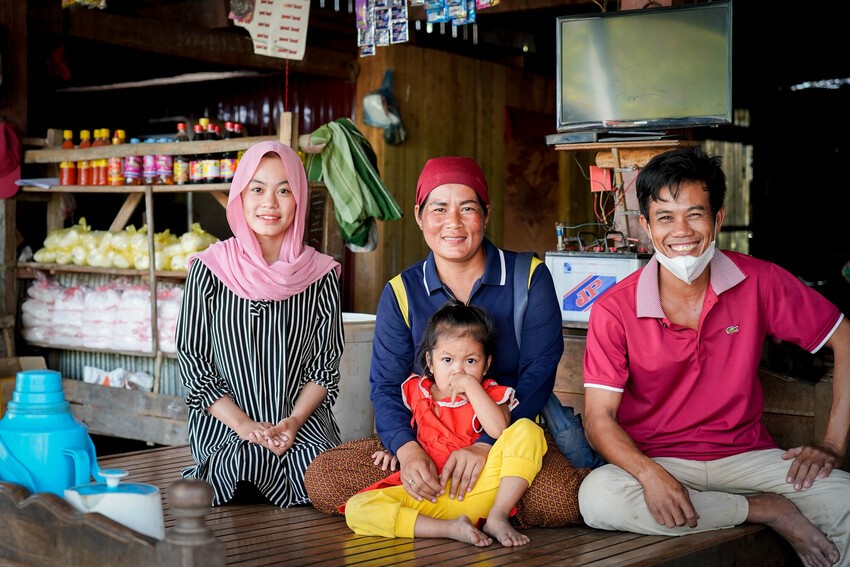 Smiling family of four sitting in their small shop, with shelves of goods, a television, and household supplies in the background.