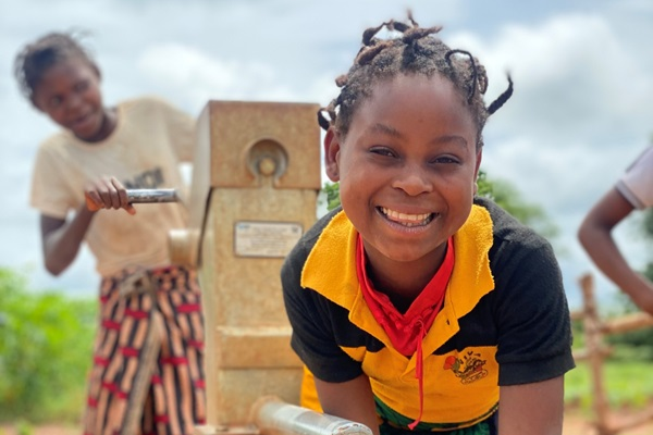 A smiling girl pours water into a bucket from a community water tap.