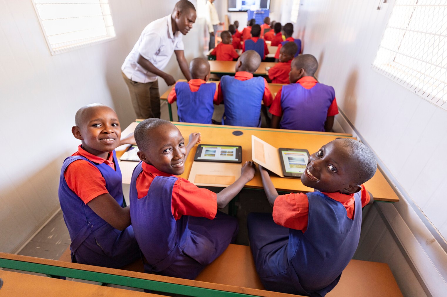 Students in red and blue uniforms using tablets at wooden desks, smiling at the camera, with a teacher assisting others in a well-lit classroom.