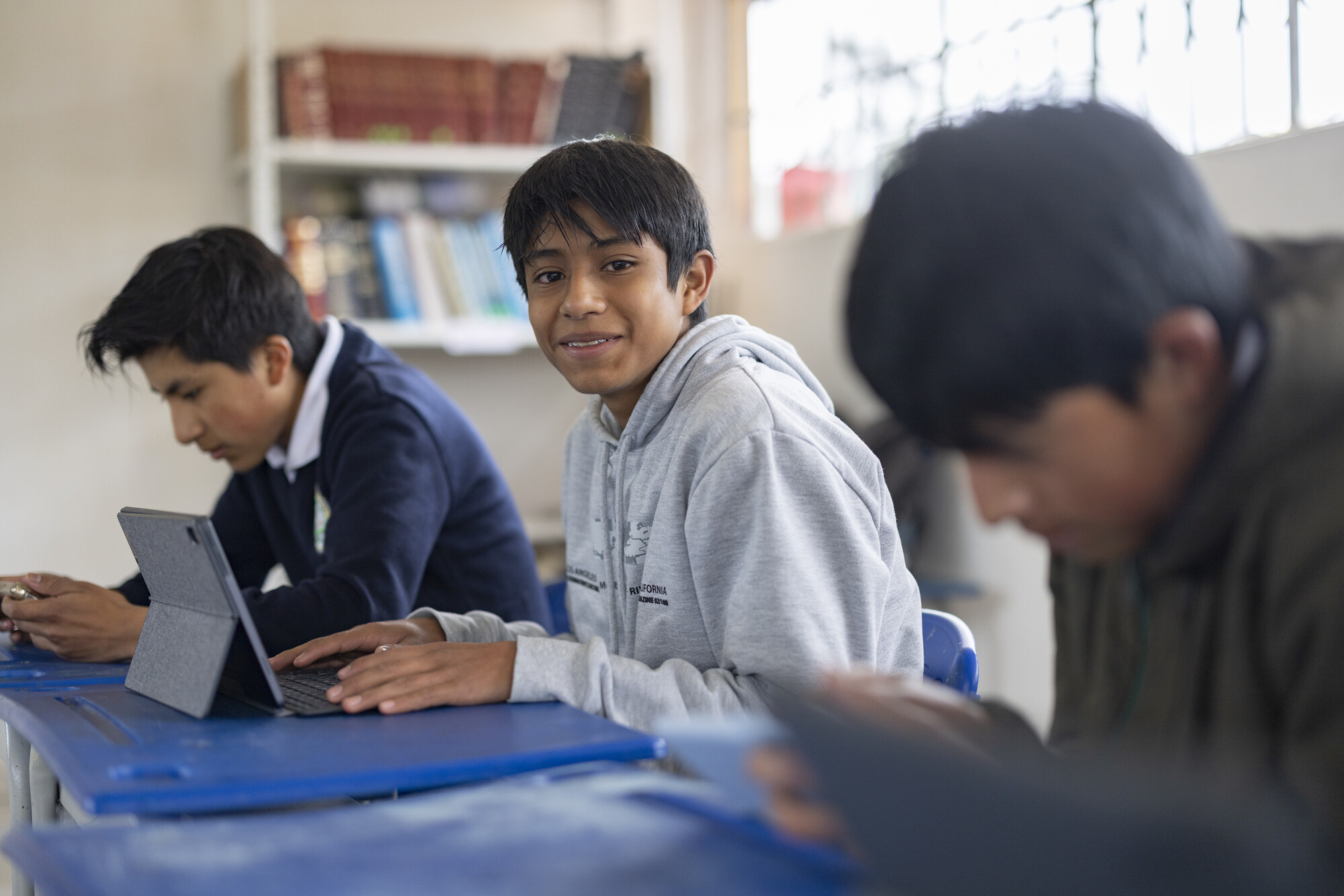 A student in a gray hoodie smiles while using a tablet in a classroom, with classmates focused on their work in the background.