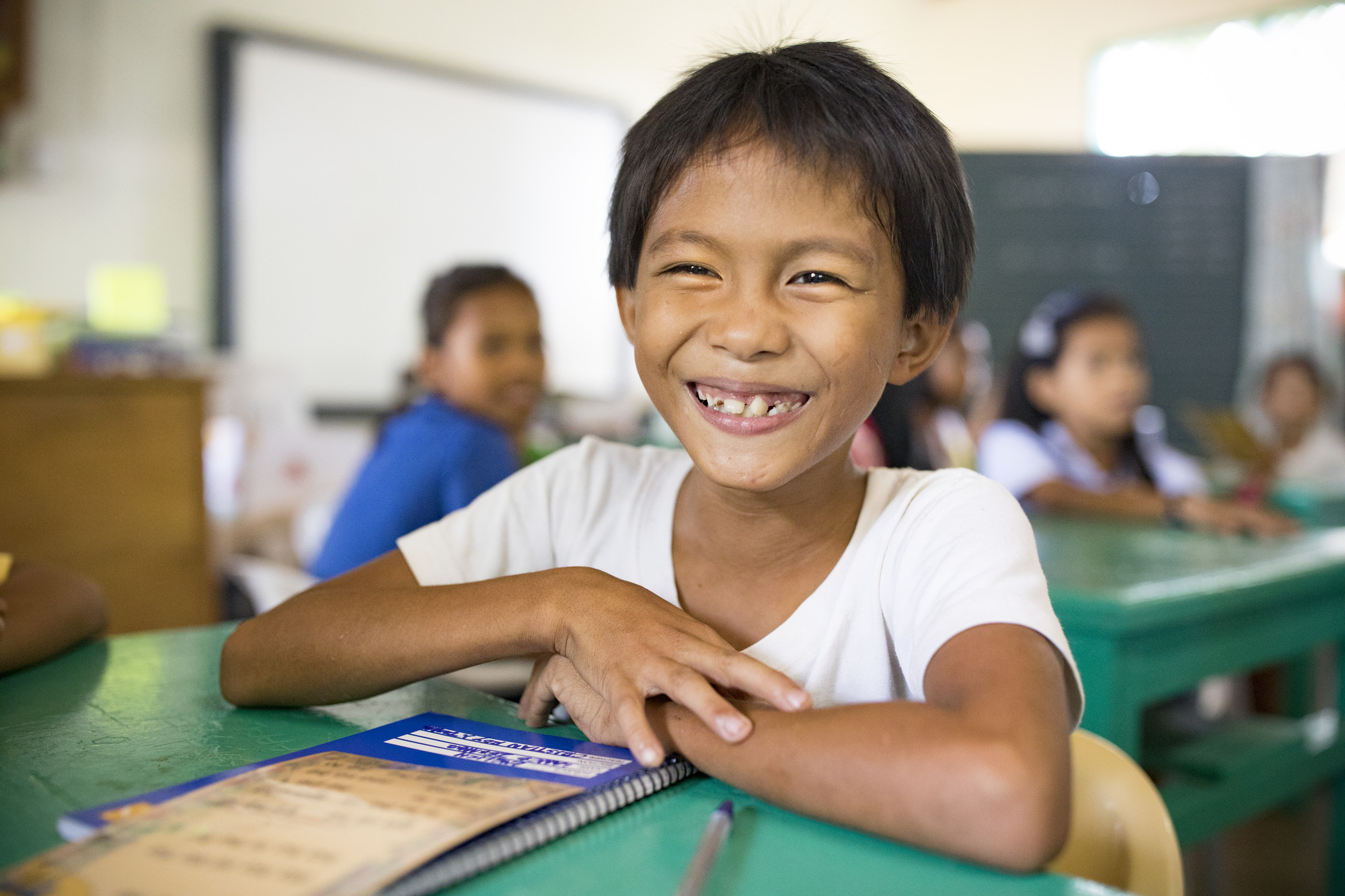 Smiling student sitting at a green desk in a classroom with other children studying in the background.