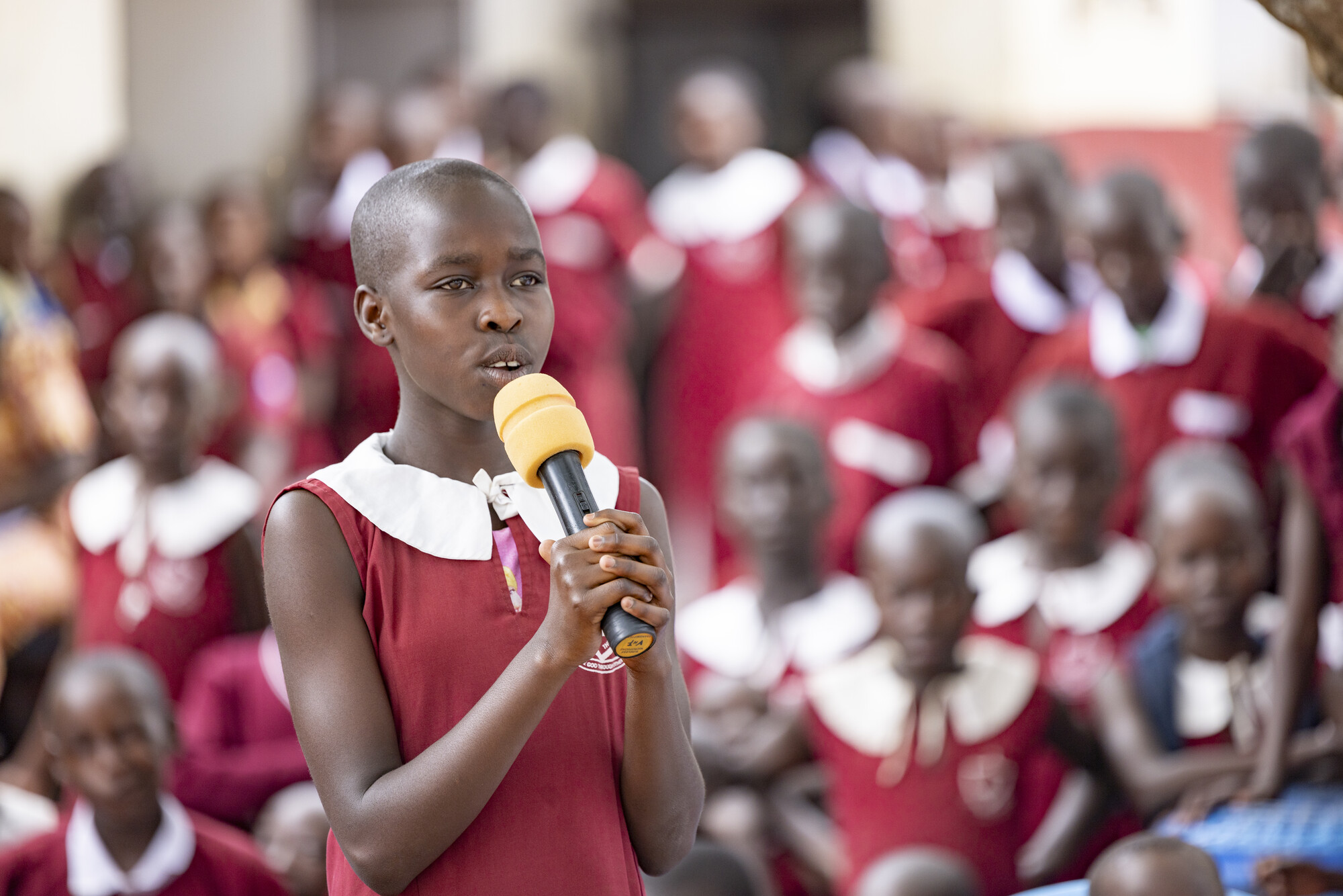 A young girl in a red and white school uniform holds a microphone and speaks to a group of students in matching uniforms in the background.