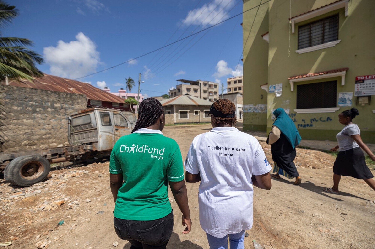 Two women wearing ChildFund Kenya and internet safety initiative shirts walk through an urban neighborhood with buildings, graffiti, and an abandoned vehicle in the background.