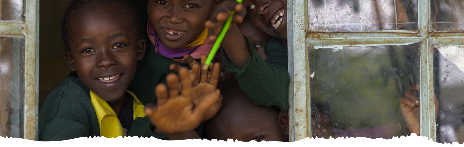 A group of children in green and yellow school uniforms smiling and waving through an old classroom window.