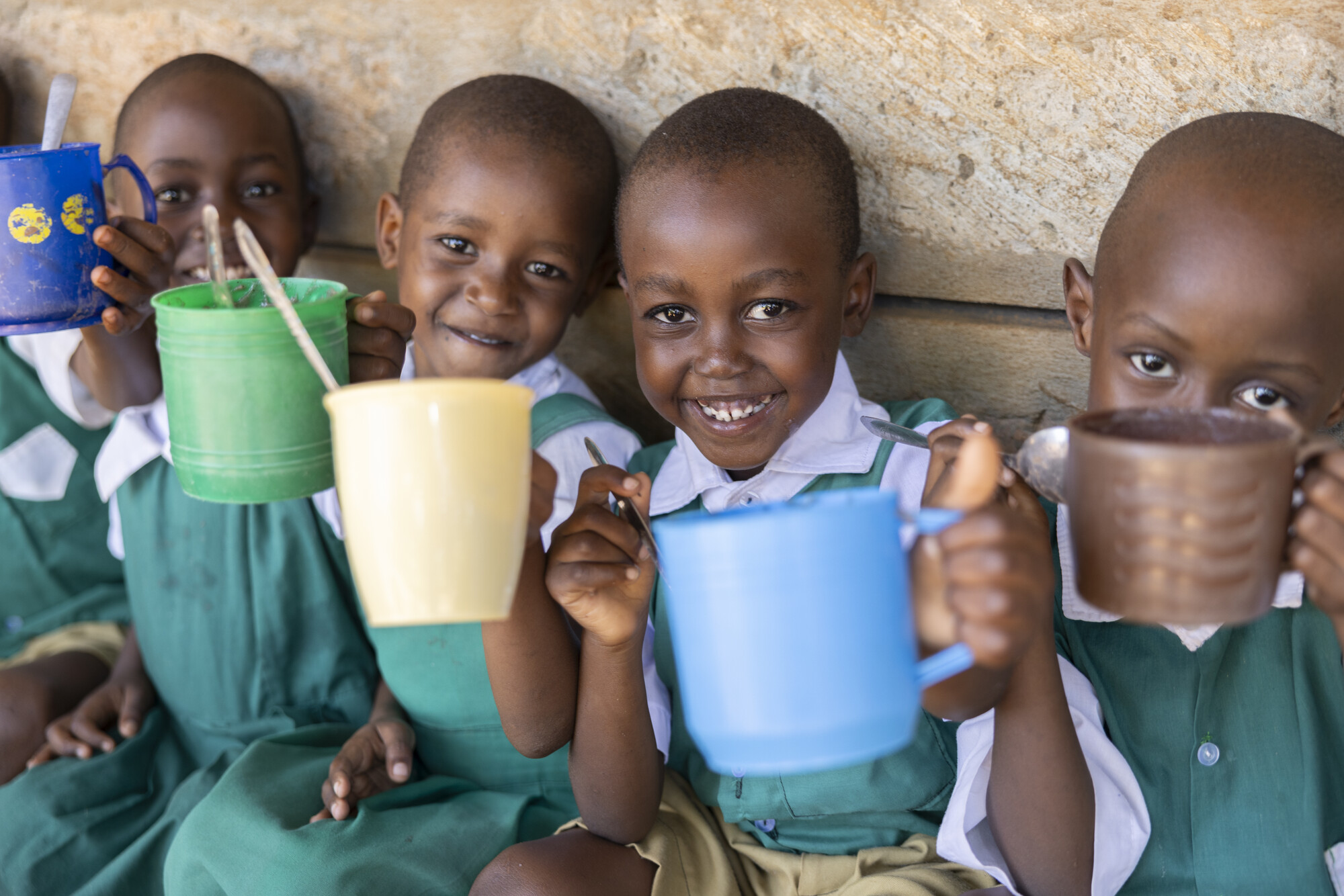Smiling schoolchildren in green and white uniforms holding colorful mugs while sitting against a stone wall.