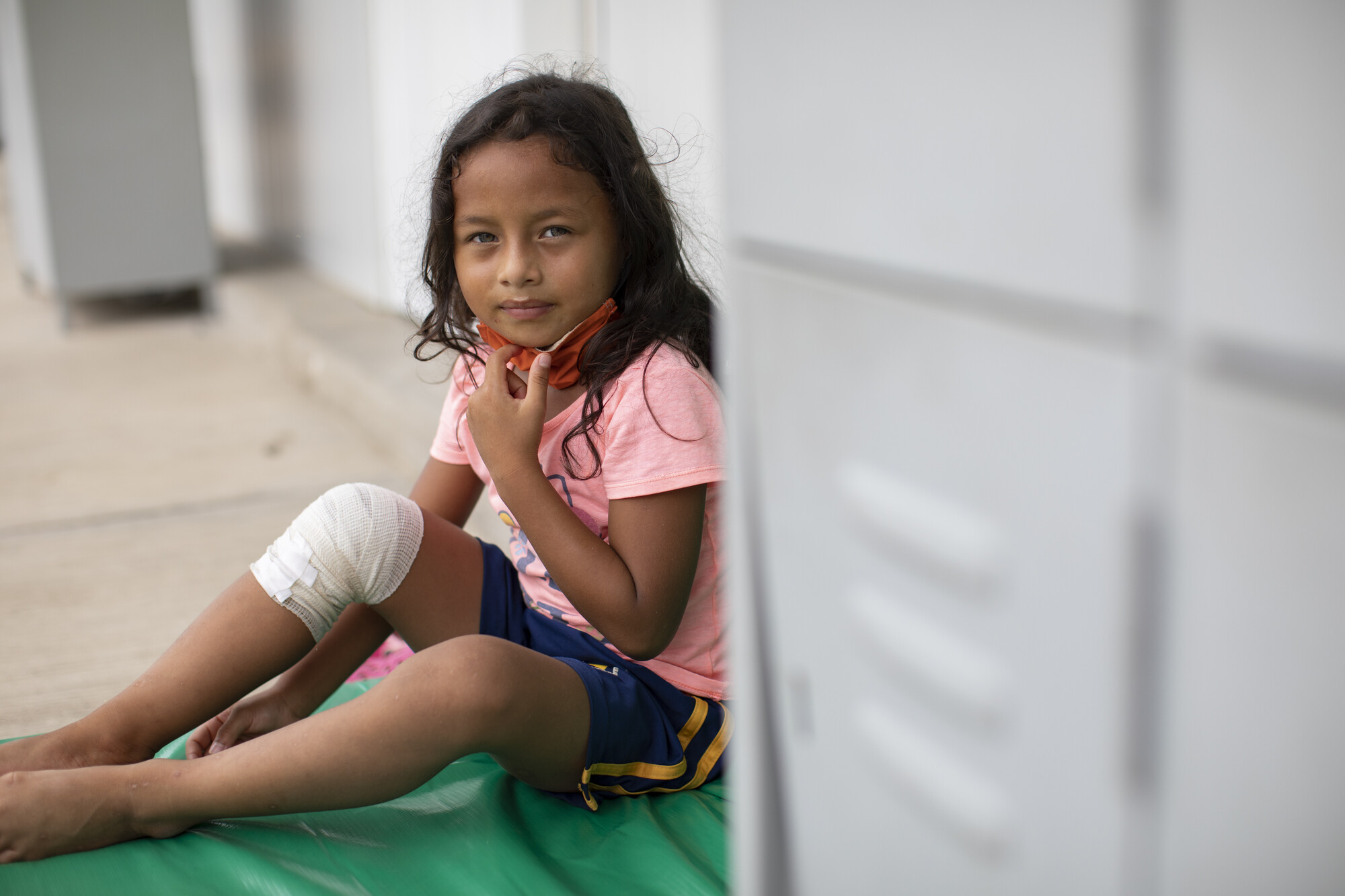 A young girl with long dark hair, wearing a pink shirt and blue shorts, sits on a mat with a bandaged knee, adjusting her mask in a temporary shelter.