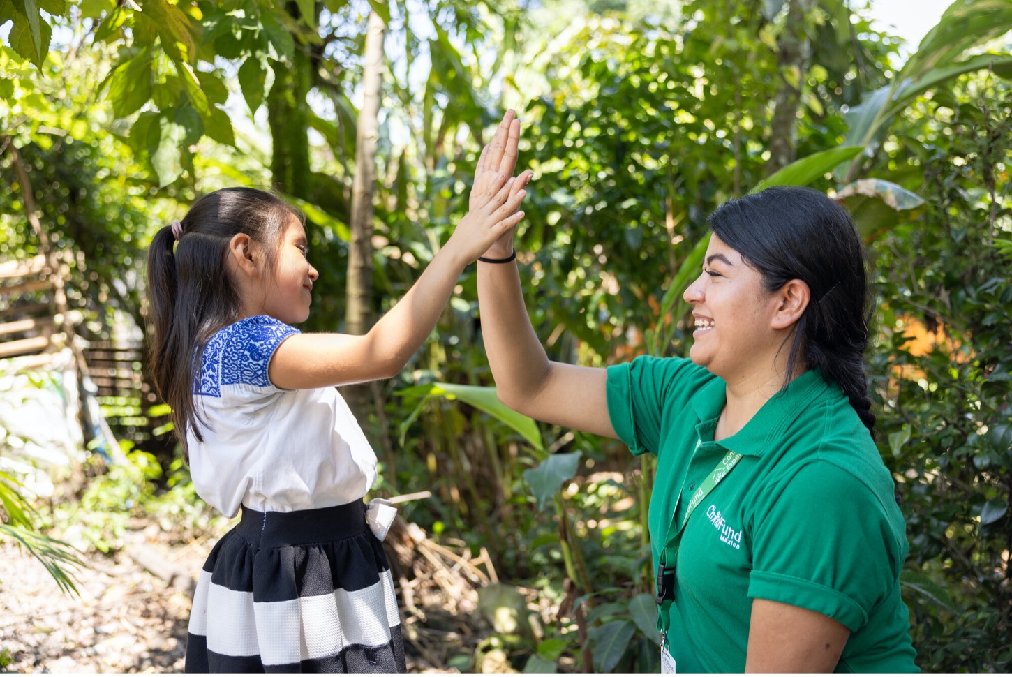 Young girl and woman in a green uniform high-fiving outdoors, surrounded by lush greenery, both smiling.