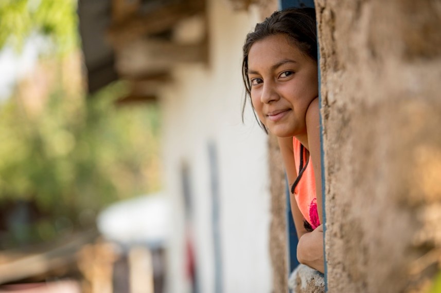 Young girl leaning out of a rustic window, smiling gently with an adobe-style building and natural background.