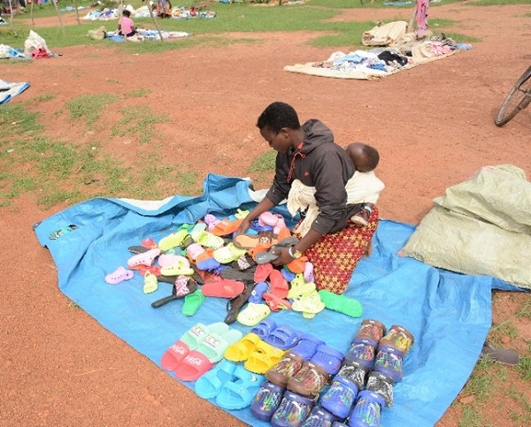 Girl sits on blanket with baby on her back at a market in Uganda, surrounded by shoes.