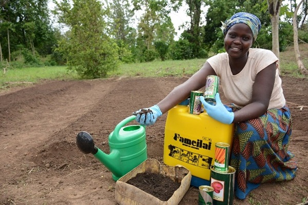 Woman crouches on ground where seedlings are growing, smiling at the camera.