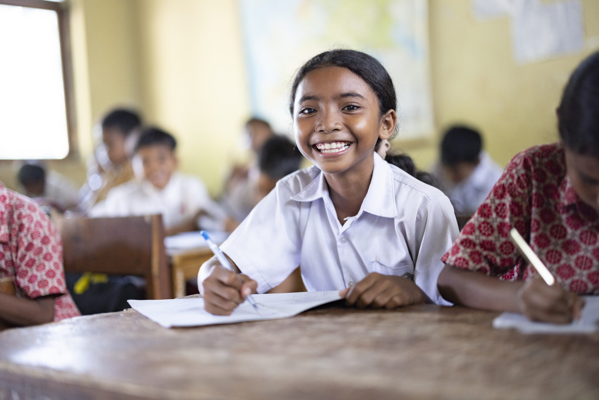 A cheerful young girl in a white school uniform writes in her notebook, surrounded by classmates in a lively classroom.