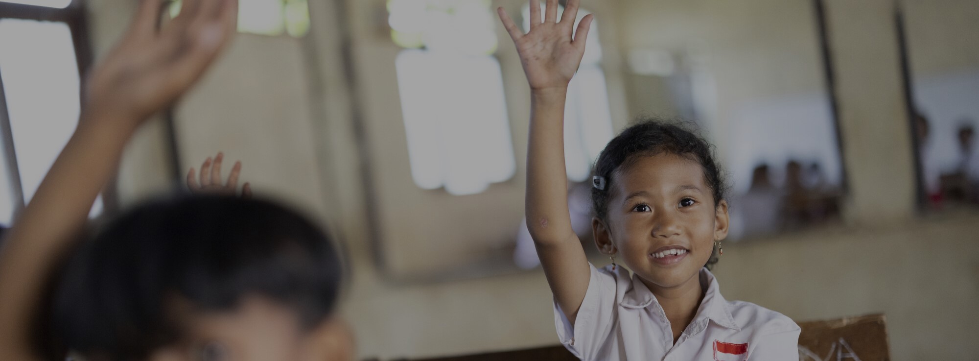 Young girl in a school uniform raising her hand in a classroom, with other students in the background and an educational message overlaid.