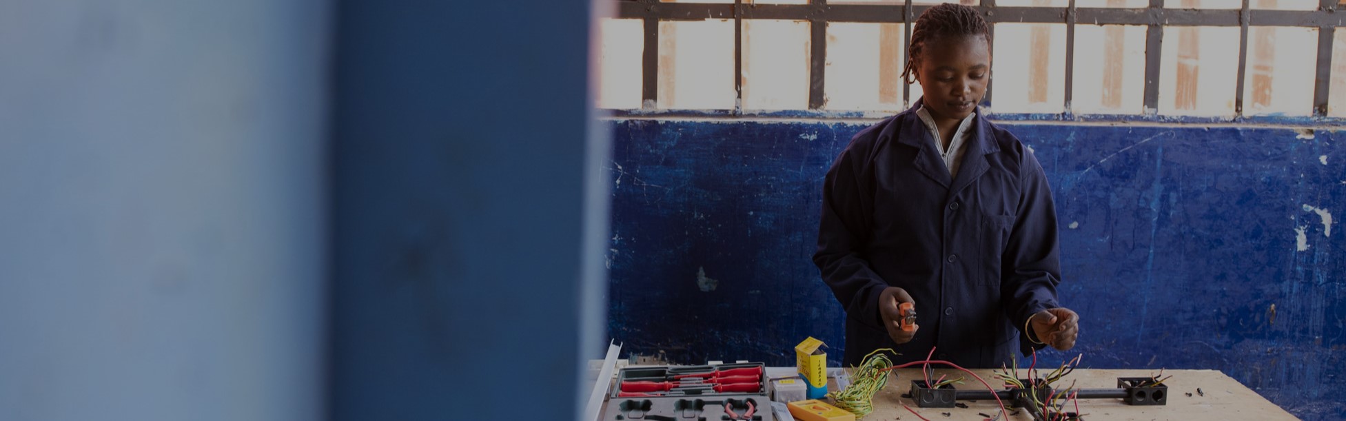 Young woman in a blue work uniform handling electrical components at a workshop table with tools, against a blue wall with a window.