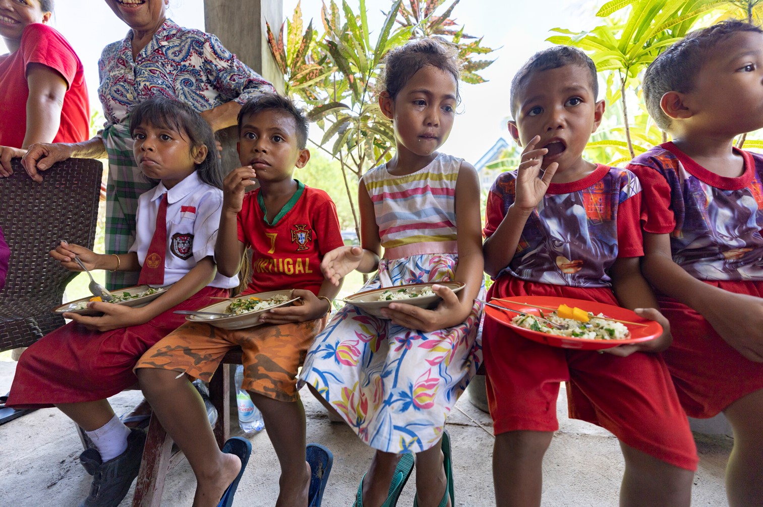 Children sitting together, eating meals from plates, dressed in colorful clothes, with smiling adults in the background in an outdoor setting.