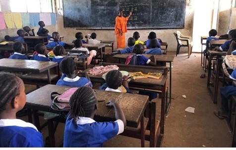 Students in blue uniforms sitting at wooden desks while a teacher writes on a blackboard in a classroom.