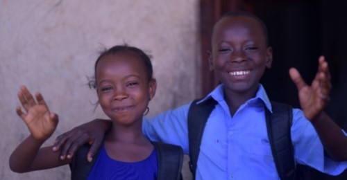 Smiling girl and boy with backpacks waving at the camera, standing in front of a textured wall.