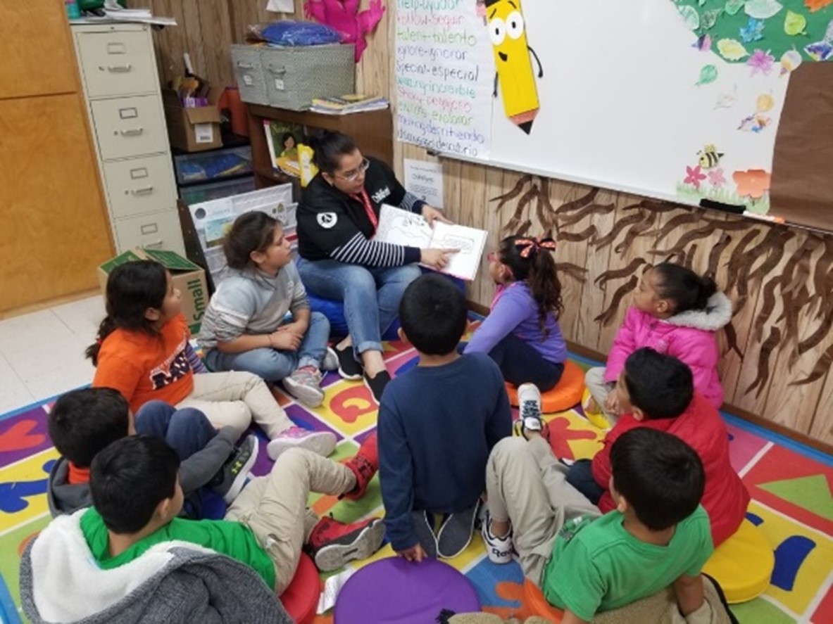 A volunteer reads a book to a group of children in the colorful corner of a classroom.