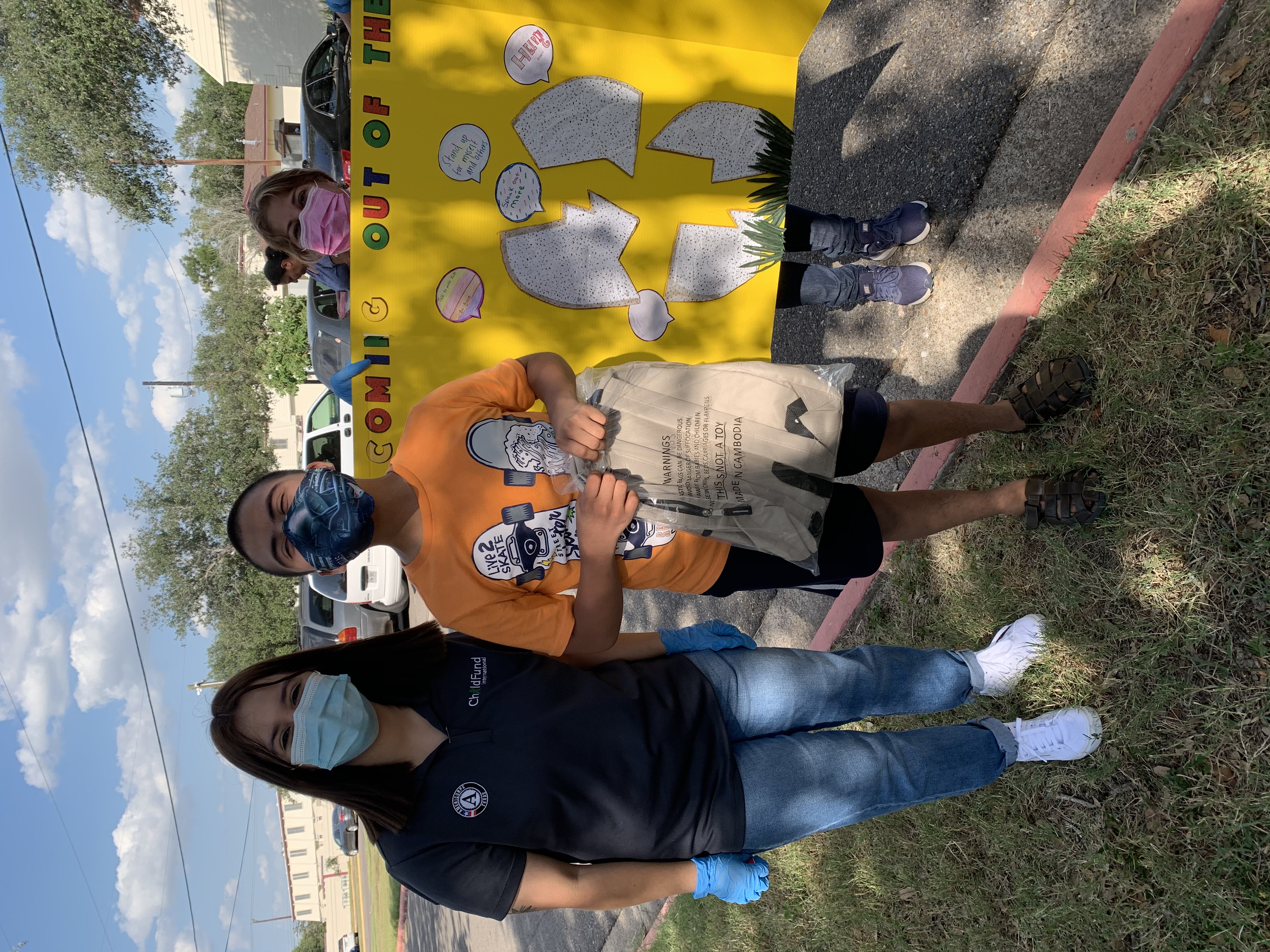 A woman in a ChildFund shirt and a teenage boy stand looking at the camera, wearing masks, in front of a piece of colorful posterboard.