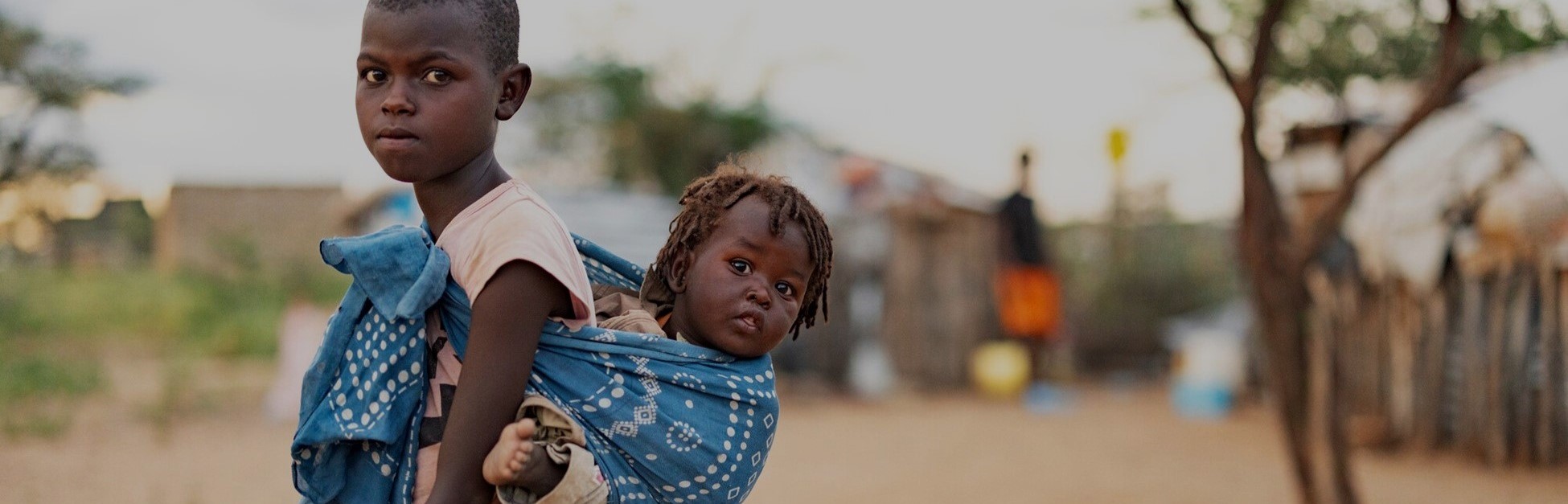 Child carrying a baby sibling in a blue cloth, walking through a rural area with simple homes and trees in the background.